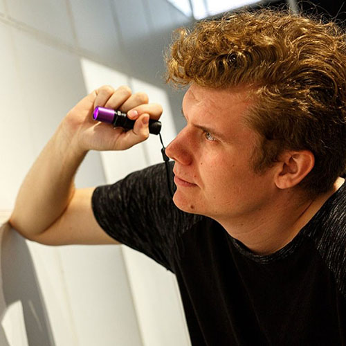 Scott is seen in profile, wearing a black t-shirt, holding a purple-tipped flashlight, looking intently ahead, in an investigative manner.